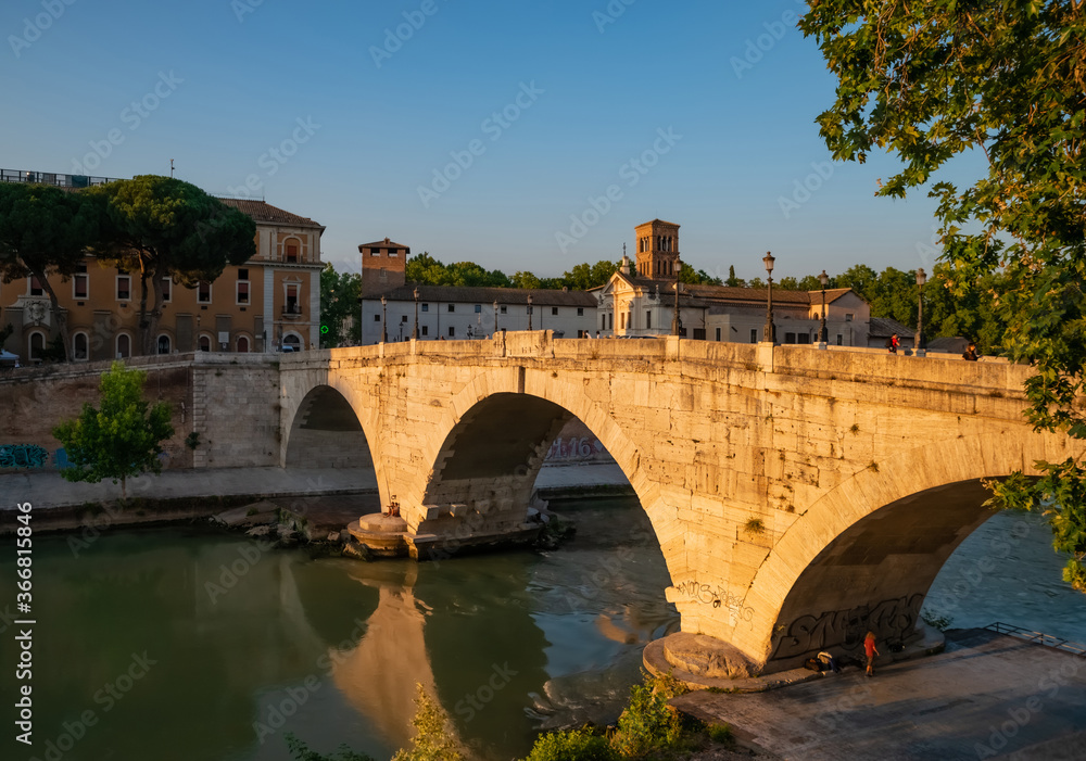 Obraz premium View of the Ponte Fabricio Rome, which connects the Tiber island to the Jewish quarter. The bridge reflects with shadows its arches in the Tevere river at sunset on a hot summer day.