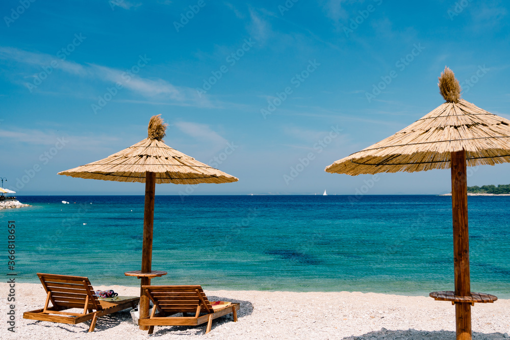 Two wooden loungers on the white sand, on a paradise beach near the town of Primosten in Croatia. Straw beach umbrella. Blue sky and azure Adriatic Sea. The concept of beach holidays and travel.