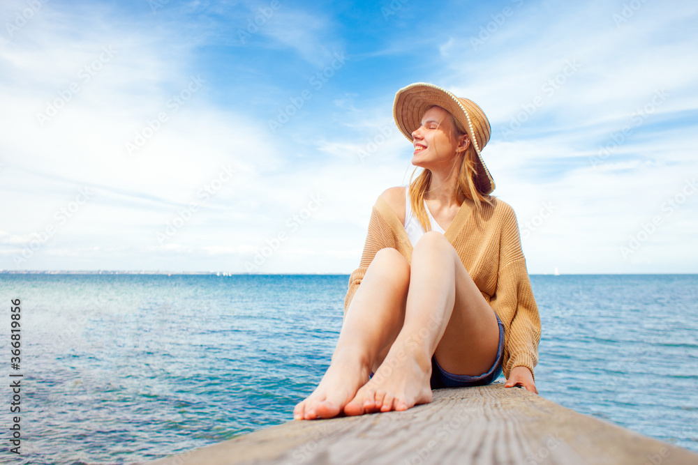 young happy girl sitting on the pier barefoot and dreaming, woman in a sun hat resting