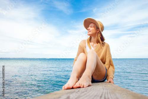 young happy girl sitting on the pier barefoot and dreaming, woman in a sun hat resting