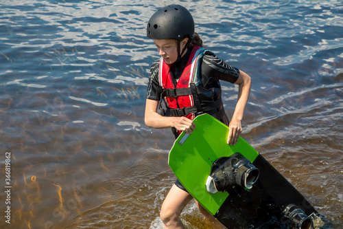 A teenage girl comes out of the water and carries Wake Board. Sports for children and teenagers. Summer pastime. Children's sports camp