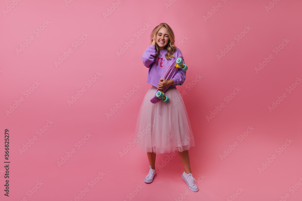 Excited charming girl in long lush skirt posing on pink background. Indoor photo of stylish female model with purple skateboard.