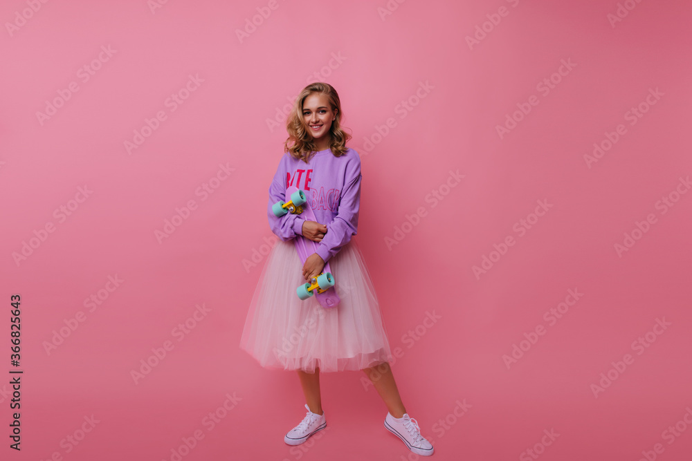 Full-length shot of glad female model with skateboard. Indoor portrait of carefree blonde girl in long lush skirt isolated on pink background. Full-length shot of glad female model with skateboard. Indoor portrait of carefree blonde girl in long lush skirt isolated on pink background.