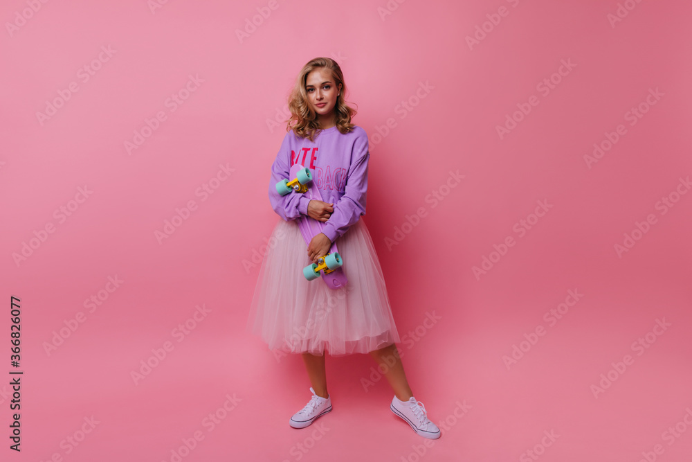 Full-length photo of enthusiastic young lady wears lush white skirt. Romantic girl with skateboard standing on rosy background. Full-length photo of enthusiastic young lady wears lush white skirt. Romantic girl with skateboard standing on rosy background.