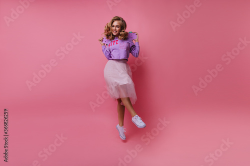 Pleasant caucasian woman in lush skirt dancing in studio. Female model in trendy clothes holding skateboard and making funny faces.
