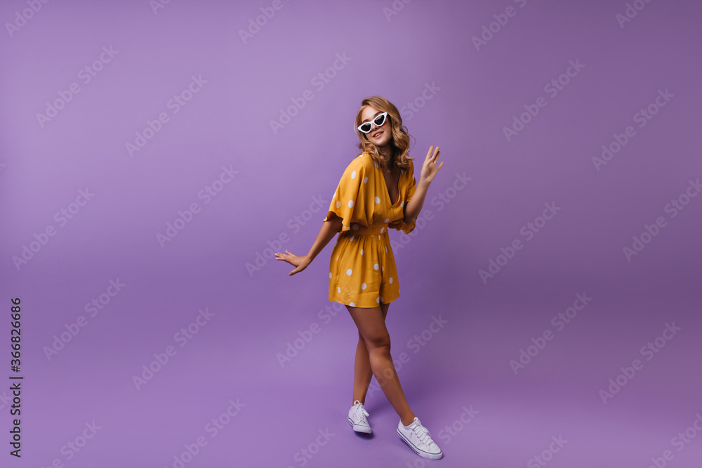 Full-length portrait of tanned gorgeous lady in white shoes. Indoor shot of romantic curly girl in yellow outfit posing on purple background.