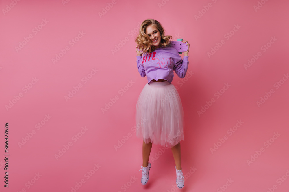 Confident girl in romantic lush skirt jumping on pink background. Pretty blonde woman with skateboard enjoying life. Confident girl in romantic lush skirt jumping on pink background. Pretty blonde woman with skateboard enjoying life.