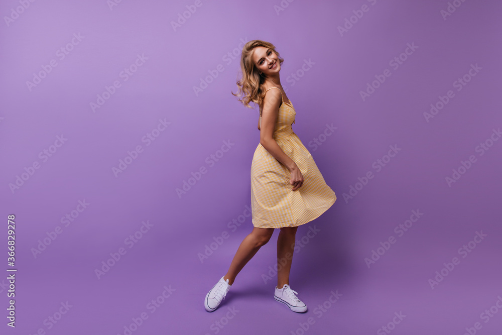 Full-length portrait of carefree female model playing with her yellow dress. Indoor photo of blithesome curly woman dancing on purple background.