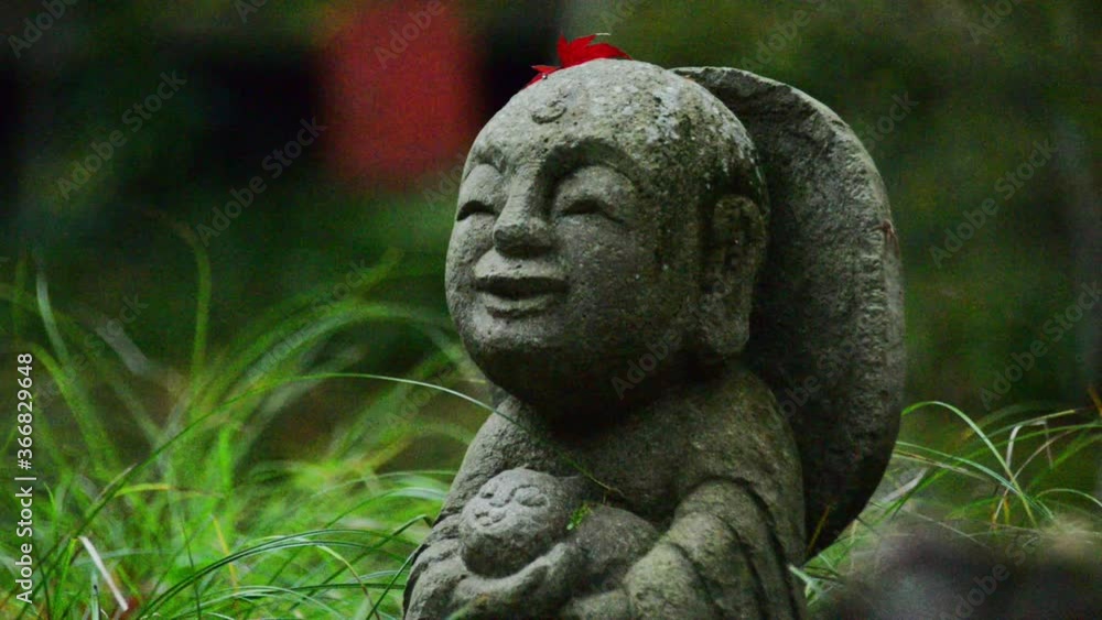 A small Jizo staute with a red leaf on its head, staying in lush green grass. Sanzen-in Buddhist temple, Ohara, Japan A small Jizo staute with a red leaf on its head, staying in lush green grass. Sanzen-in Buddhist temple, Ohara, Japan