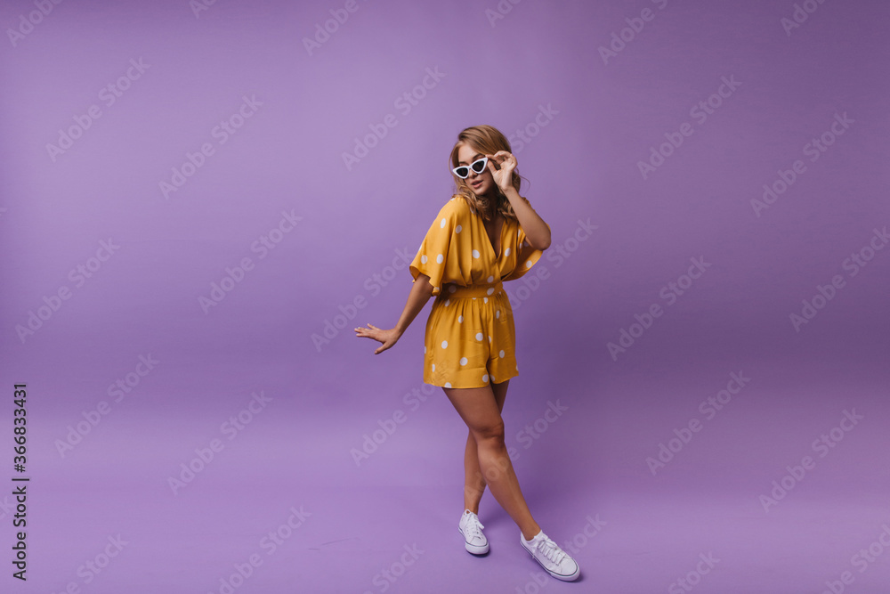 Indoor photo of amazing fair-haired lady dancing on purple background. Dreamy european woman in yellow dress and sunglasses having fun in studio.