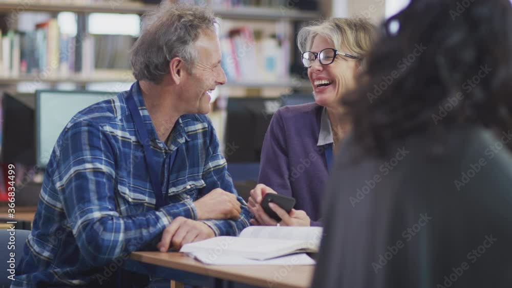 Female teacher helping mature male student studying and using mobile phone in library learning IT skills in evening class - shot in slow motion Female teacher helping mature male student studying and using mobile phone in library learning IT skills in evening class - shot in slow motion