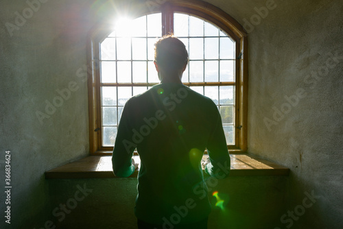 Rear view of young man looking through closed wooden vintage window with sunlight streaming in