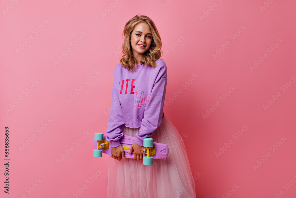 Interested blonde girl with curly hairstyle posing with longboard. Refined white woman in purple shirt standing on pink background. Interested blonde girl with curly hairstyle posing with longboard. Refined white woman in purple shirt standing on pink background.
