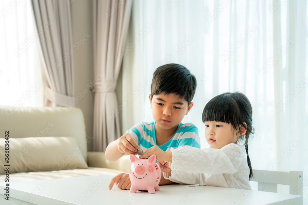 Two Asian cute brother and sister insert coin in piggy bank together on the table in living room at home. Concept of bonding of sibling, friendship and saving, investment money concept Two Asian cute brother and sister insert coin in piggy bank together on the table in living room at home. Concept of bonding of sibling, friendship and saving, investment money concept