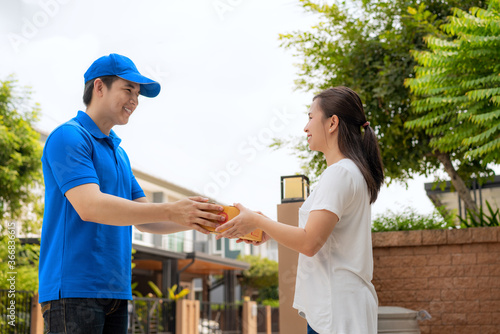Asian delivery young man in blue uniform smile and holding a cardboard boxes in front house and Asian woman accepting a delivery of boxes from deliveryman. Advertising, Transportation Concept. © ake1150