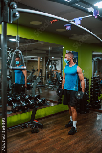 A young muscular caucasian athlete man with a mask on his face exercises in the gym and lifts weights in front of a mirror. COVID 19 coronavirus protection