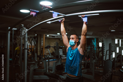 Muscular young fitness man with medical mask doing exercises on horizontal bar in a gym club. COVID 19 coronavirus protection © Dragica