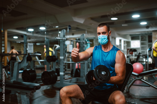 A young muscular caucasian athlete with a mask on his face, with a weight in his hand gives a thumb up in the gym. Protection against COVID - 19 coronavirus