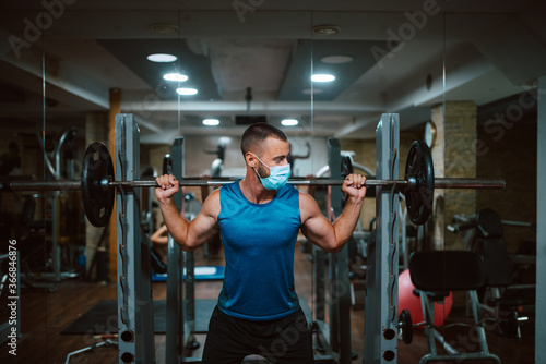 A young caucasian athlete man with a mask on his face exercises and lifts weights in the gym. COVID 19 coronavirus protection