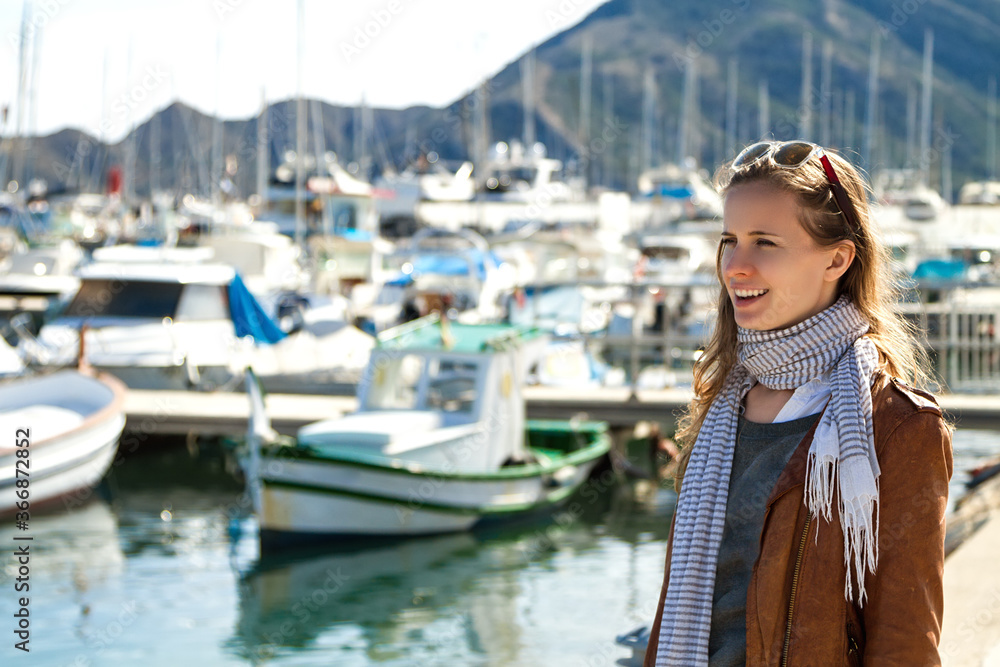 Portrait of a modern fashionable beautiful woman with long hair and in sunglasses, brown jacket, scarf smiling and looking forward on the background of a sunny beach, coastline and a view of the seasc