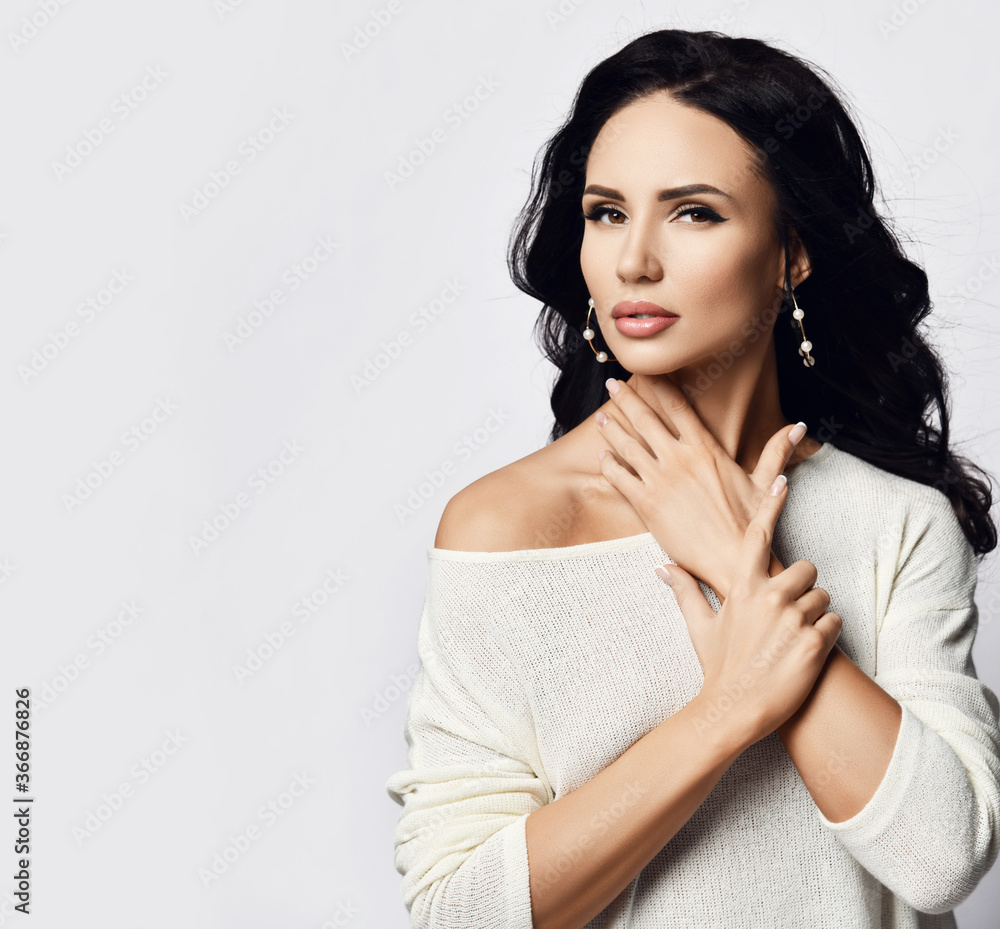 Young noble brunette woman with long hair in one-shoulder sweater and stylish earrings stands with hand at her neck Young noble brunette woman with long hair in one-shoulder sweater and stylish earrings stands with hand at her neck