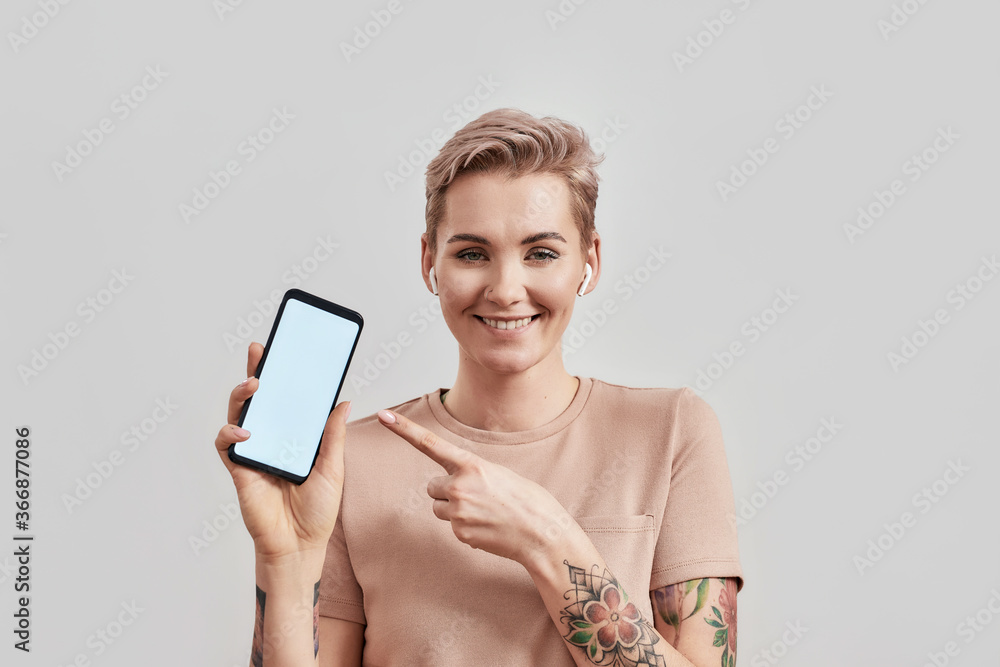 Portrait of tattooed woman with pierced nose and short hair in wireless earbuds or earphones smiling at camera, pointing at smartphone with blank screen isolated over light background Portrait of tattooed woman with pierced nose and short hair in wireless earbuds or earphones smiling at camera, pointing at smartphone with blank screen isolated over light background