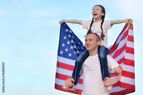 Happy daughter sits on her father's shoulders and holds the American flag above her head. American Independence Day. Happy future concept. Freedom. Election.Copy space for text.