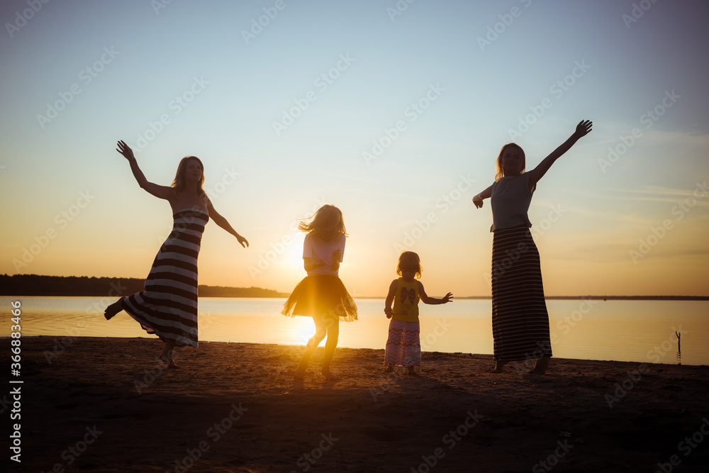 Fototapeta premium Silhouettes of children and their mothers jumping and having fun on the beach in sunset light. Good mood and pastime among the younger and older generation.