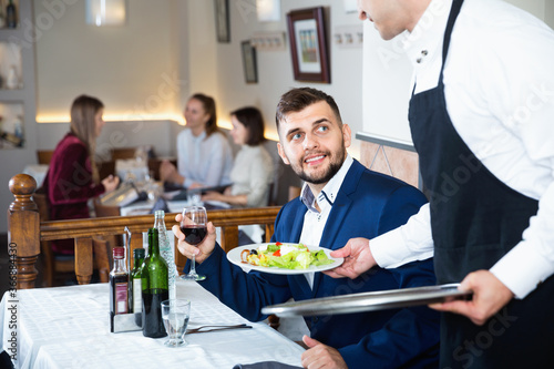 Glad cheerful positive waiter serving delicious salads to handsome young man at restaurant