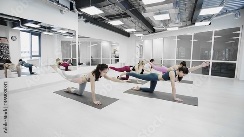 Sporty attractive girls are doing stretching exercises forward bending head to knee position while sitting on colorful yoga mats in light wellness studio.