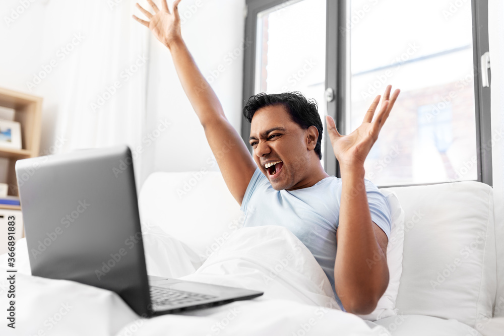 people, technology and success concept - happy smiling indian man with laptop computer lying in bed and making winning gesture at home people, technology and success concept - happy smiling indian man with laptop computer lying in bed and making winning gesture at home