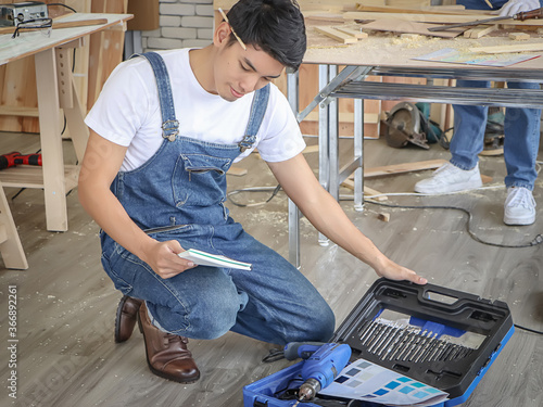 Young and handsome Asian repairman sitting  in indoor  workshop , reading instructiom with drill box in front of him . photo