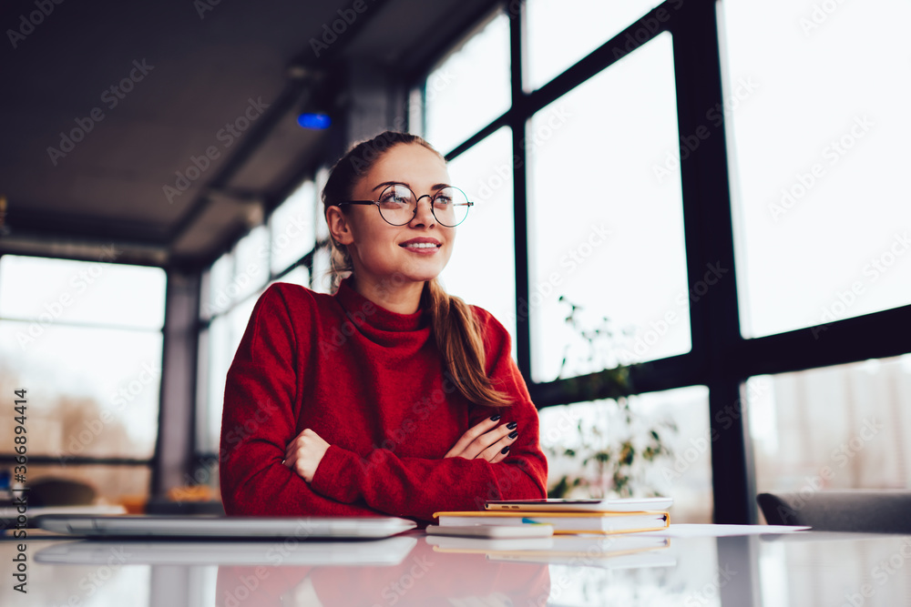 Fototapeta premium Young attractive blonde female in stylish eyeglasses dreaming whine sitting in loft interior place.Charming positive hipster girl dressed in red sweater looking away and thinking during recreating