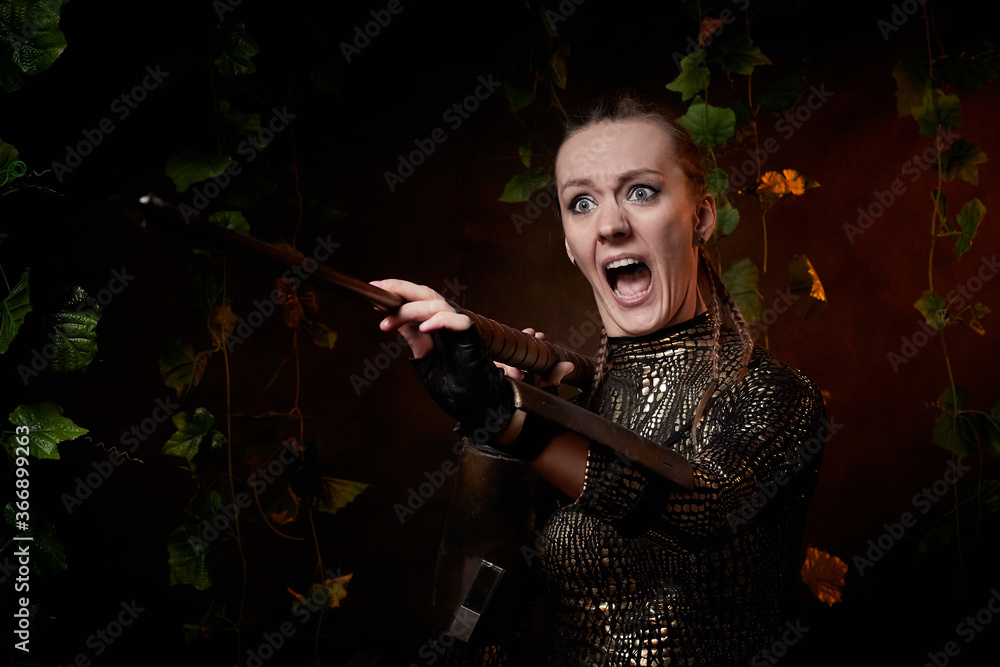 Valkyrie girl in shiny military armor and with a spear in a dark room with plants and vines. Model during a photo shoot, the actress during the shooting of the film.