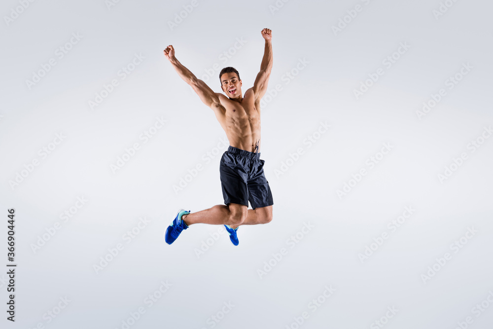 Full size photo of handsome sportsman dark skin guy naked chest jump high up celebrate competition winning raise arms up wear shorts sneakers isolated white color background Full size photo of handsome sportsman dark skin guy naked chest jump high up celebrate competition winning raise arms up wear shorts sneakers isolated white color background