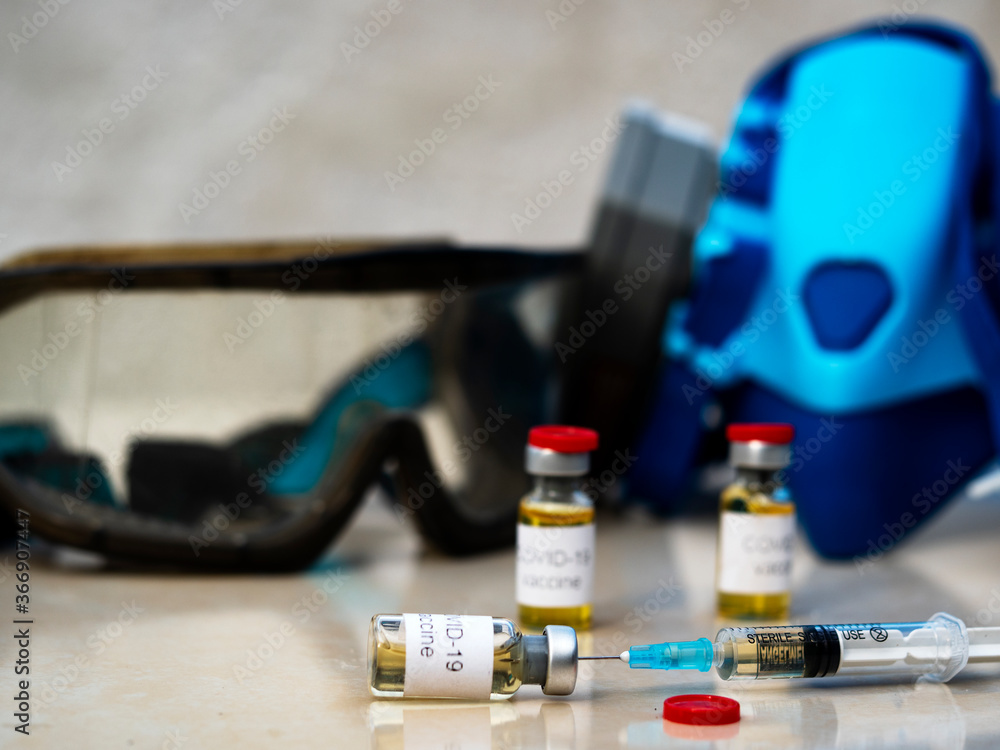 In this photo illustration, vials labelled as Coronavirus vaccine and a syringe against the background of a medical respirator and goggles seen displayed as the world anxiously