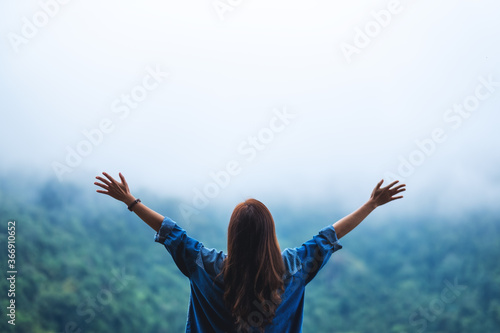 Rear view image of a female traveler open arms and looking at a beautiful green mountain on foggy day