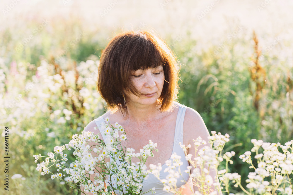 Middle aged mature red haired woman picking wild daisy flowers in field. Elderly female in a meadow and pick flower bouquet on summer day. Spending time outdoors in spring. Middle aged mature red haired woman picking wild daisy flowers in field. Elderly female in a meadow and pick flower bouquet on summer day. Spending time outdoors in spring.