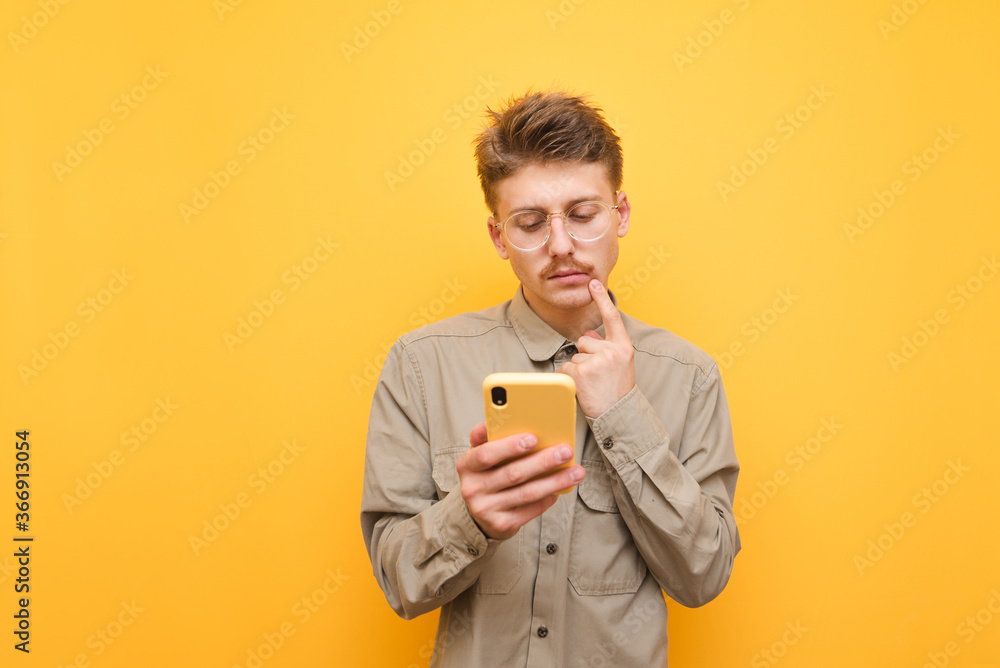Thoughtful young man with mustache and glasses uses a smartphone against a yellow background,looks at the screen and thinks,wearing a shirt.Funny nerd with a serious face is using the phone. Isolated Thoughtful young man with mustache and glasses uses a smartphone against a yellow background,looks at the screen and thinks,wearing a shirt.Funny nerd with a serious face is using the phone. Isolated