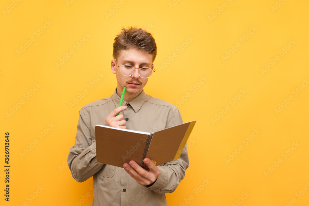 Portrait of serious student with notepad and pen in hand, looking intently at notebook and thinking about task. Nerd in shirt and glasses with pen and notepad isolated on yellow background. Copy space Portrait of serious student with notepad and pen in hand, looking intently at notebook and thinking about task. Nerd in shirt and glasses with pen and notepad isolated on yellow background. Copy space