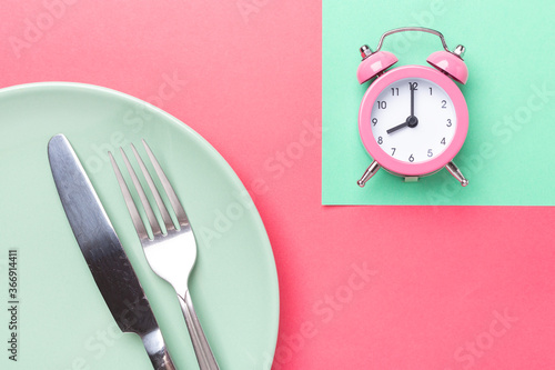 Pink alarm clock, fork, knife and empty plate on colored paper background. Intermittent fasting concept