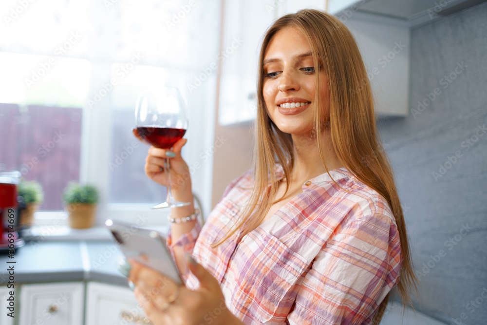 Happy relaxed young woman standing in kitchen with glass of red wine and using her smartphone Happy relaxed young woman standing in kitchen with glass of red wine and using her smartphone