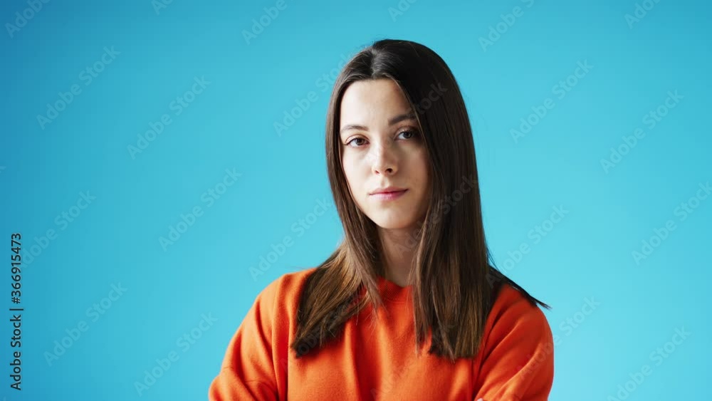 Studio portrait of confident young woman looking into camera with neutral expression and folded arms against blue background - shot in slow motion Studio portrait of confident young woman looking into camera with neutral expression and folded arms against blue background - shot in slow motion