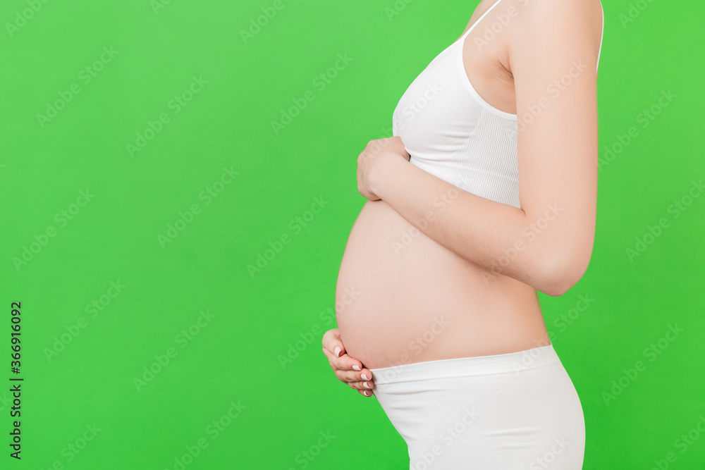 Cropped image of pregnant woman in white underwear touching her belly at green background. Future parent is expecting her child. Copy space Cropped image of pregnant woman in white underwear touching her belly at green background. Future parent is expecting her child. Copy space