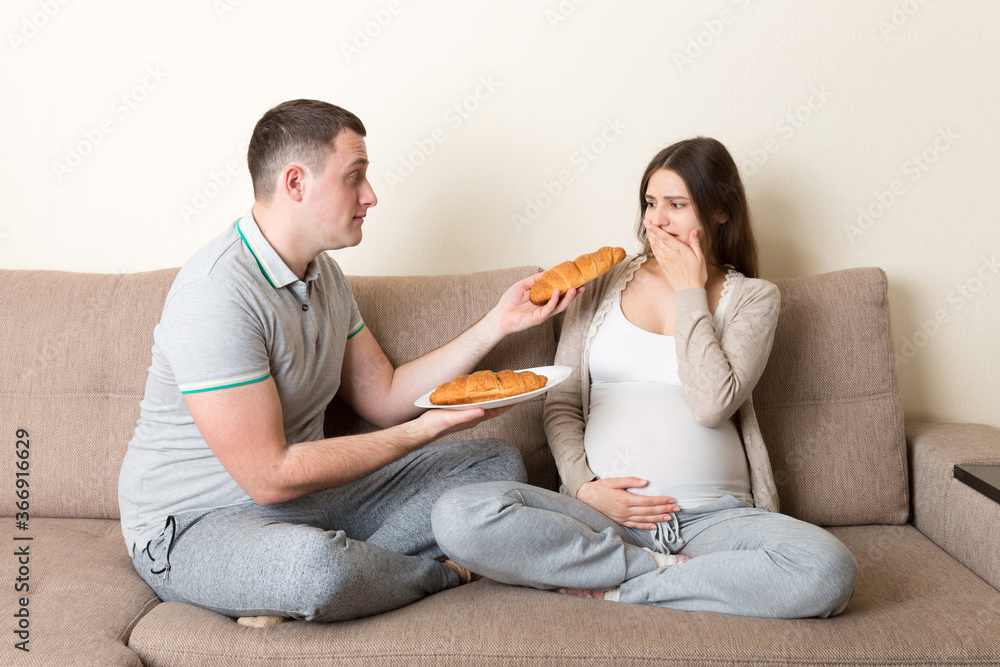 Husband offers croissants to his pregnant wife but she refuses and makes stop gesture because she feels sick. Feeling bad during pregnancy concept Husband offers croissants to his pregnant wife but she refuses and makes stop gesture because she feels sick. Feeling bad during pregnancy concept