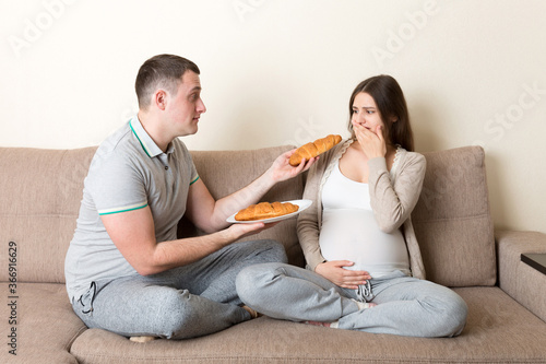Husband offers croissants to his pregnant wife but she refuses and makes stop gesture because she feels sick. Feeling bad during pregnancy concept