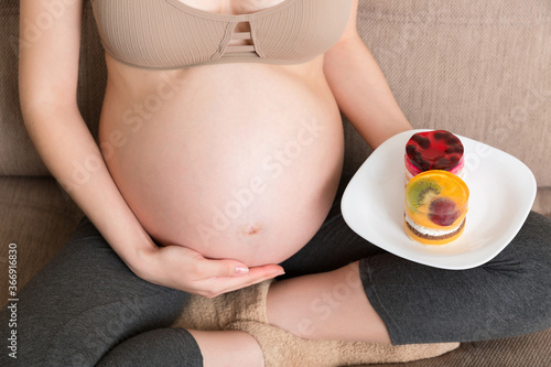 Close up of hungry pregnant woman is eating tasty desserts sitting on the sofa at home. Enjoying sweet food during pregnancy concept