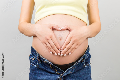 Close up of pregnant woman in opened jeans making heart on her belly at colorful background with copy space. Love to a future baby concept © sosiukin