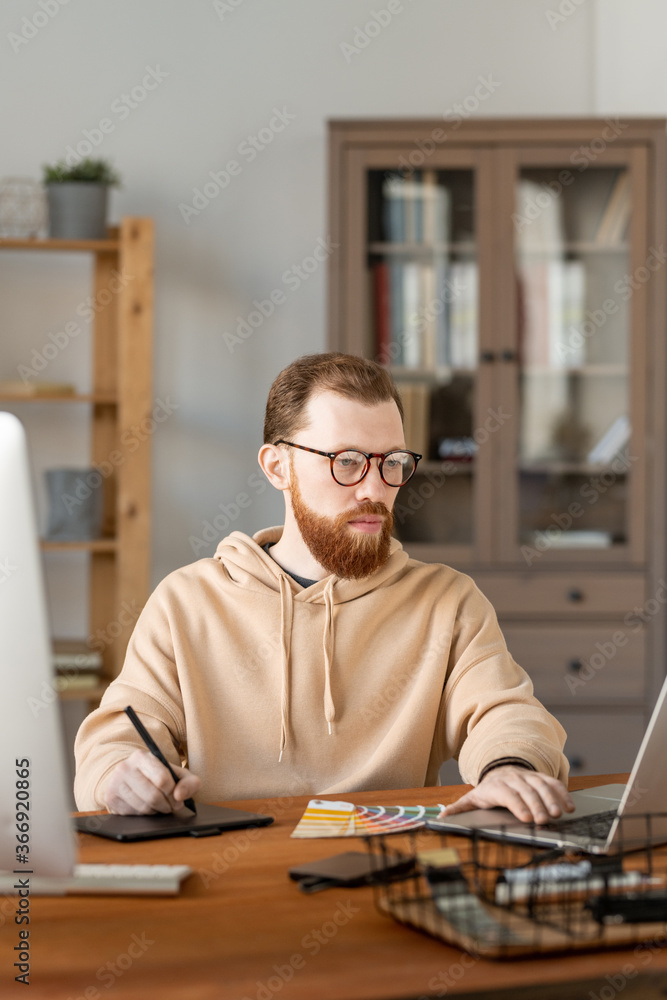 Fototapeta premium Serious young bearded man in eyeglasses drawing design sketch on graphics tablet and editing it on laptop