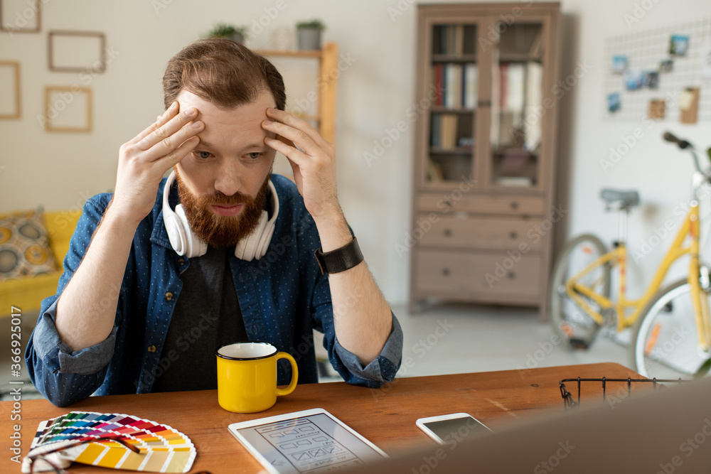 Puzzled young UI designer with beard sitting at table and holding head in hands while thinking of interface layout Puzzled young UI designer with beard sitting at table and holding head in hands while thinking of interface layout